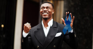 A triumphant black man in a suit, holding up a prostate cancer awareness ribbon and smiling after beating colon cancer.