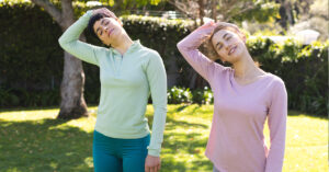 Happy biracial lesbian couple practicing yoga standing and stretching necks in sunny garden