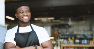 Cheerful entrepreneur in an apron, standing with crossed arms, exuding confidence and positivity in a successful restaurant.