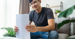 Content asian man sorts and reviews printed trial entry forms while sitting on his couch in front of his laptop,